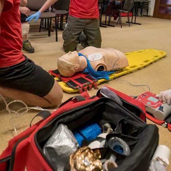 UETDREL006-feature-2 Two individuals in red shirts practice CPR on a rescue dummy lying on a yellow stretcher, with medical equipment nearby.