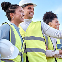 Three construction workers in safety vests and helmets discuss plans on a site, set against a clear blue sky.