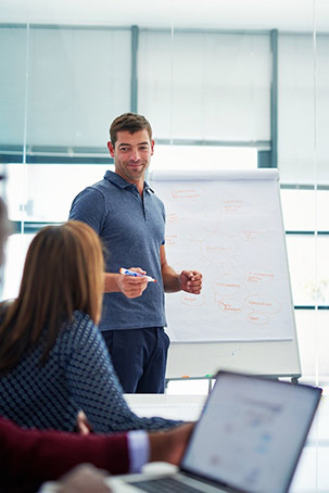 A man presents in a meeting, standing beside a whiteboard, while students attentively listen from their seats with laptops.