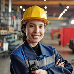 A worker in a yellow hard hat and blue jacket stands confidently in a warehouse, holding a walkie-talkie amidst industrial surroundings.