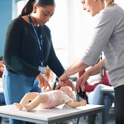 Two women demonstrate infant CPR on a baby doll in a classroom setting, with students observing in the background.