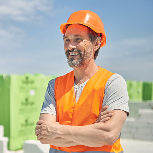 A construction worker in a bright orange vest and hard hat stands confidently on a building site with green materials in the background.