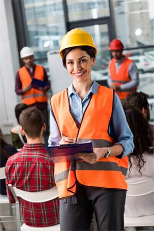 A woman in a yellow hard hat and orange safety vest holds a clipboard while overseeing a training session with an audience.