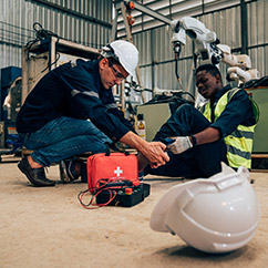 A worker in a hard hat administers first aid to a colleague seated on the ground, with a first aid kit and safety equipment nearby.