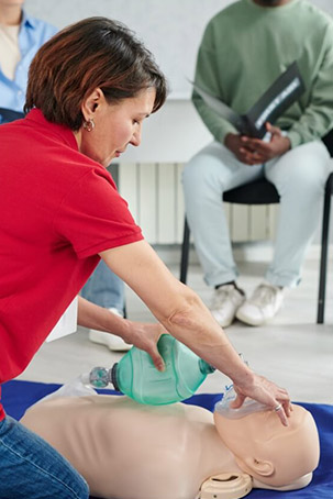 A person in a red shirt performs CPR on a training mannequin while others observe, highlighting a lifesaving skills workshop.